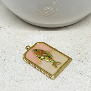 May include: A rectangular gold-toned pendant with a colorful fish design. The fish features a gold body, a rainbow-colored fin, and a black eye. The pendant has a pink background and a gold border, suitable for jewelry making.