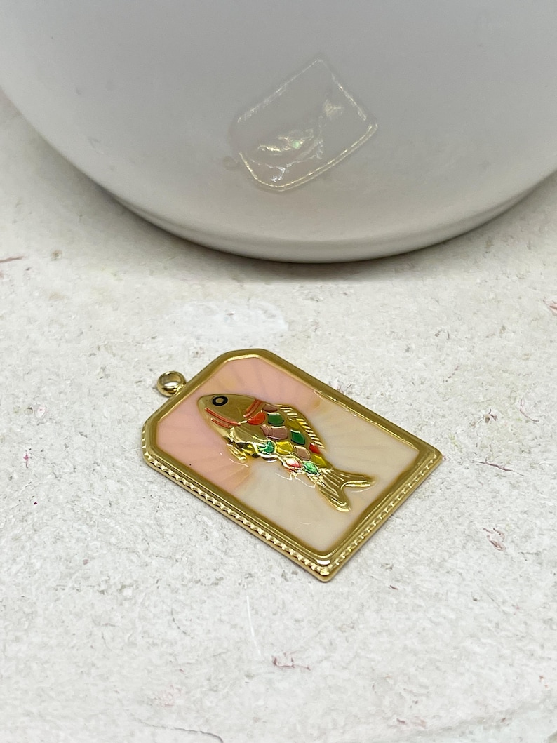 May include: A rectangular gold-toned pendant with a colorful fish design. The fish features a gold body, a rainbow-colored fin, and a black eye. The pendant has a pink background and a gold border, suitable for jewelry making.