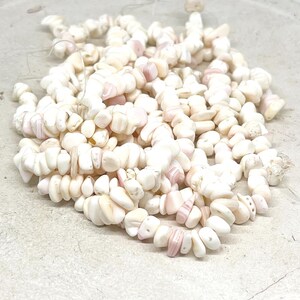 Könnte beinhalten: Ein Haufen kleiner, unregelmäßiger, cremefarbener Perlen mit zarten rosa Farbtönen. Die Perlen, möglicherweise aus Muscheln oder Stein, sind auf einer hellen Oberfläche verstreut. Diese Perlen könnten zur Schmuckherstellung verwendet werden.