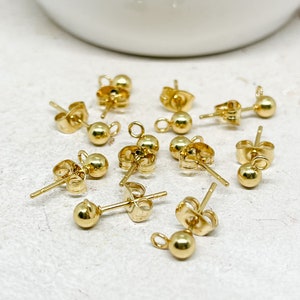 May include: A collection of small, gold-colored ball stud earring components. Each earring post has a small ball at the end and a butterfly clutch backing. The earrings are scattered on a white surface.