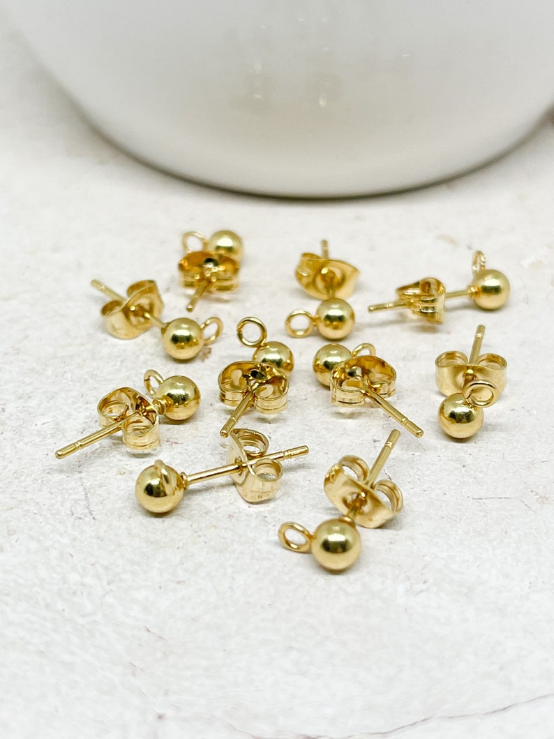 May include: A collection of small, gold-colored ball stud earring components. Each earring post has a small ball at the end and a butterfly clutch backing. The earrings are scattered on a white surface.