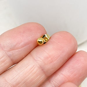 May include: A close-up of a gold-colored earring back. The earring back has a small, round ball on one end and a flat, butterfly-shaped clasp on the other. The earring back is held between fingers.