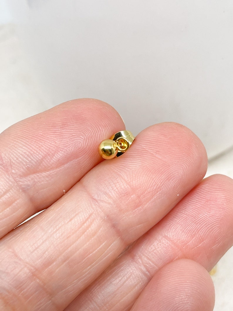 May include: A close-up of a gold-colored earring back. The earring back has a small, round ball on one end and a flat, butterfly-shaped clasp on the other. The earring back is held between fingers.