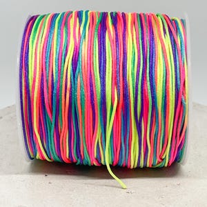 Könnte beinhalten: Eine Spule mit buntem Regenbogen-Gummiband. Das Band besteht aus mehreren Fäden in Neonrosa, Grün, Blau, Gelb und Lila.
