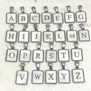 Op de afbeelding: Een collectie van zesentwintig rechthoekige metalen hangers, elk voorzien van een witte parelmoeren inleg met een zwarte hoofdletter van A tot Z in het midden. Elke hanger heeft een klein metalen oogje aan de bovenkant om op te hangen.