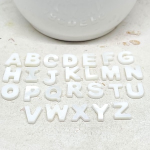 Könnte beinhalten: Eine Sammlung weißer, perlmuttartiger Buchstaben auf einer hellen Oberfläche. Die Buchstaben sind in einer Serifenschriftart gehalten und scheinen einzelne Bastelstücke zu sein. Im Hintergrund befindet sich ein weißes, abgerundetes Objekt.