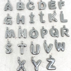 May include: A collection of silver-colored alphabet letter charms, each with a small loop at the top for attaching to a chain or bracelet. The letters are in a rounded, bubble-like font, and are arranged in rows.