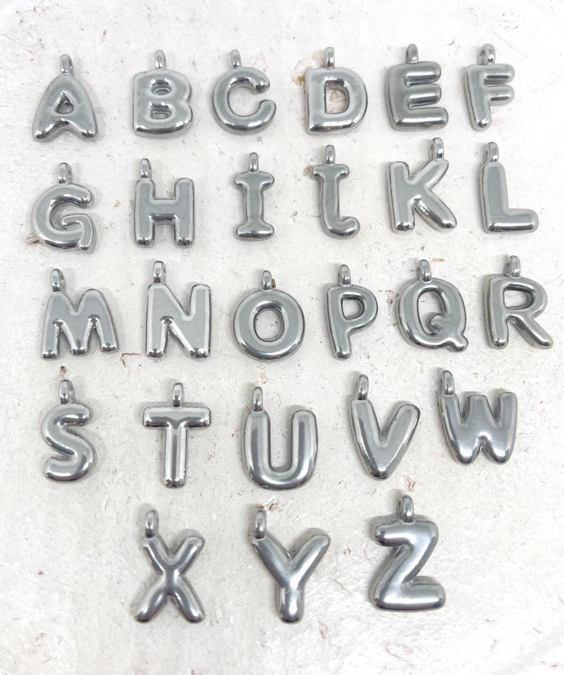May include: A collection of silver-colored alphabet letter charms, each with a small loop at the top for attaching to a chain or bracelet. The letters are in a rounded, bubble-like font, and are arranged in rows.