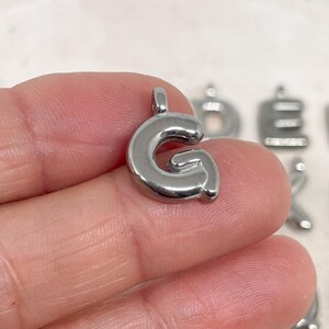 May include: A shiny, silver-colored metal charm in the shape of the letter "G". The charm has a rounded, bubble-like design and a small loop at the top for attaching to a chain or bracelet. Other letter charms are visible in the background.