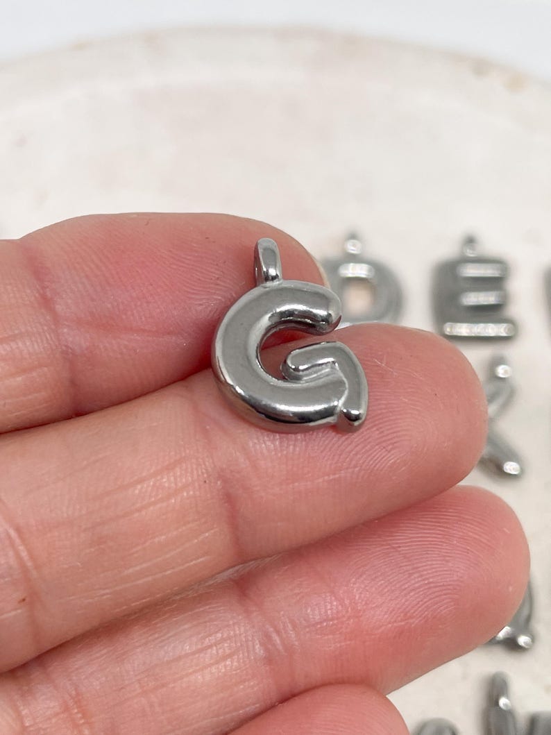May include: A shiny, silver-colored metal charm in the shape of the letter "G". The charm has a rounded, bubble-like design and a small loop at the top for attaching to a chain or bracelet. Other letter charms are visible in the background.