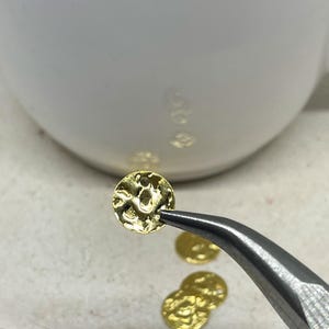 May include: A close-up shot shows a small, textured, gold-coloured disc held by tweezers. Several other similar discs are visible, along with a white ceramic object in the background. The discs appear to be jewellery components.