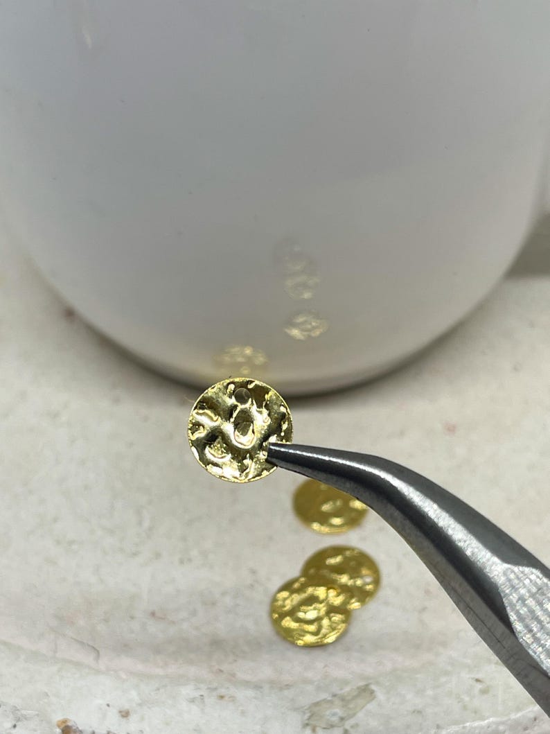 May include: A close-up shot shows a small, textured, gold-coloured disc held by tweezers. Several other similar discs are visible, along with a white ceramic object in the background. The discs appear to be jewellery components.