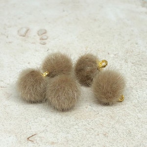 May include: Four brown faux fur pom poms with gold metal loops. The pom poms are soft and fluffy.