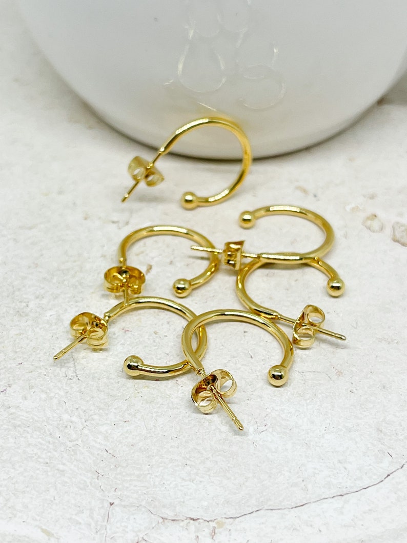 May include: Four gold hoop earrings with a small ball at the end of each hoop. The earrings are on a white surface.