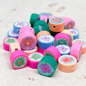 Könnte beinhalten: Eine Sammlung bunter runder Perlen mit Schmetterlingsmotiven. Die Perlen sind aus Polymer Clay gefertigt und in verschiedenen Farben erhältlich, darunter Pink, Grün, Blau und Orange. Jede Perle zeigt ein anderes Schmetterlingsdesign mit filigranen Details.