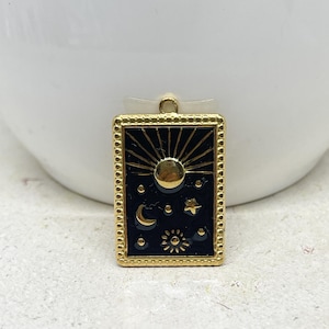 Puede incluir: Un colgante rectangular dorado con un fondo negro con dise&ntilde;os celestiales. El colgante tiene un sol radiante, luna, estrellas y sol. El colgante est&aacute; enmarcado con un borde de cuentas y tiene un peque&ntilde;o lazo en la parte superior para colgar.