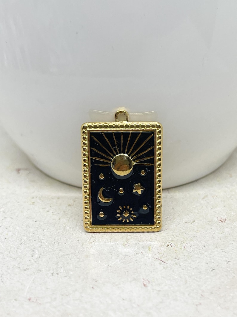 Puede incluir: Un colgante rectangular dorado con un fondo negro con dise&ntilde;os celestiales. El colgante tiene un sol radiante, luna, estrellas y sol. El colgante est&aacute; enmarcado con un borde de cuentas y tiene un peque&ntilde;o lazo en la parte superior para colgar.