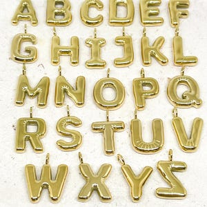 Op de afbeelding: Een verzameling goudkleurige alfabetletterbedels, elk met een lus om aan een ketting of armband te bevestigen. De letters zijn in een vet, afgerond lettertype en zijn gerangschikt in rijen en kolommen.