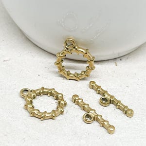 Könnte beinhalten: Vier goldfarbene Schmuckverschlüsse. Zwei sind kreisförmig mit einem strukturierten Design, und zwei sind stangenförmig mit einer Schlaufe an einem Ende. Die Verschlüsse befinden sich auf einer weißen Oberfläche mit unscharfem Hintergrund.