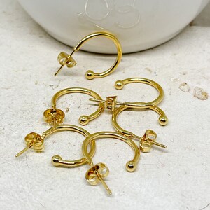 May include: Four gold hoop earrings with a small ball at the end of each hoop. The earrings are on a white surface.