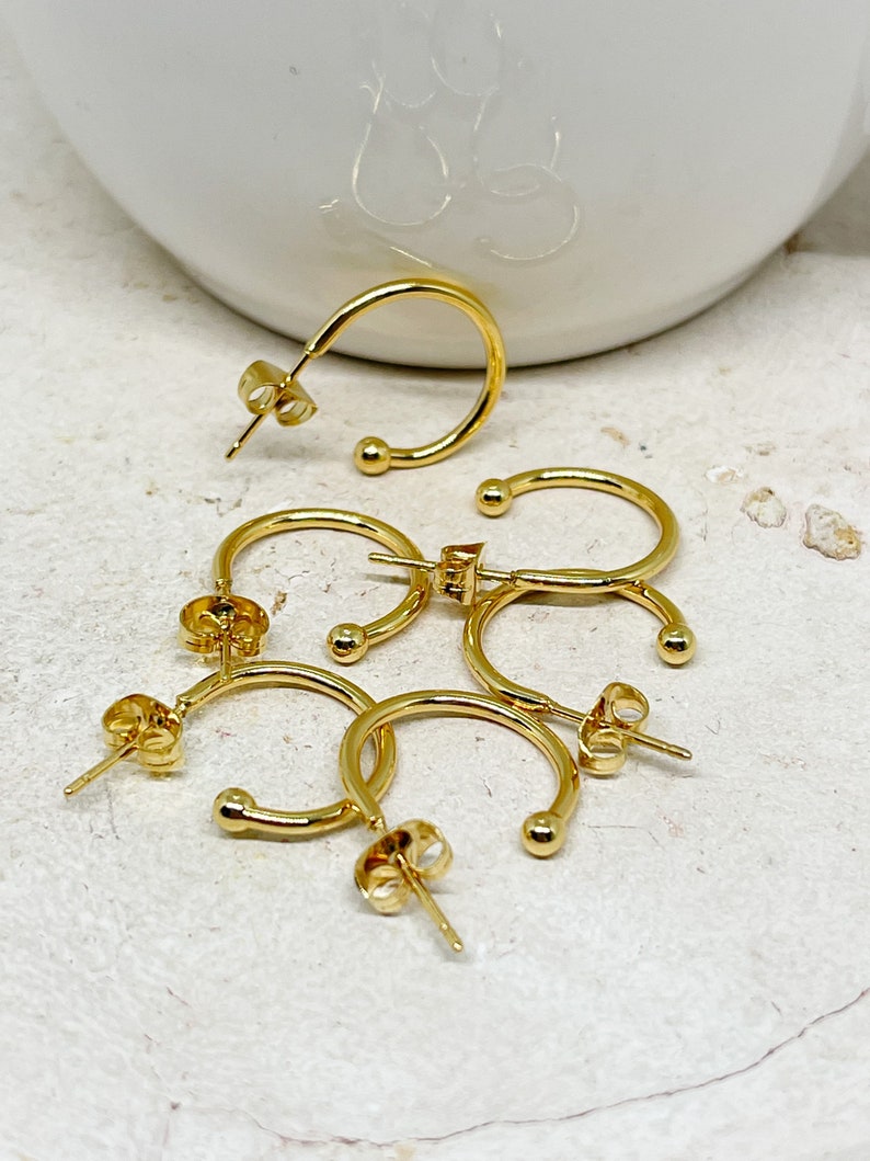May include: Four gold hoop earrings with a small ball at the end of each hoop. The earrings are on a white surface.