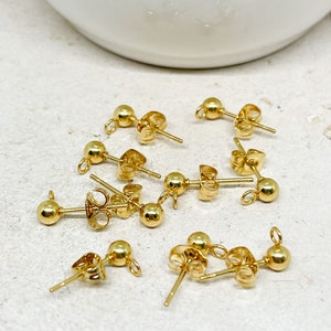 May include: A collection of gold-colored ball stud earring components. Each component features a small, round ball at the front, a post for piercing the ear, and a loop for attaching additional elements. The earrings are displayed on a light-colored surface.