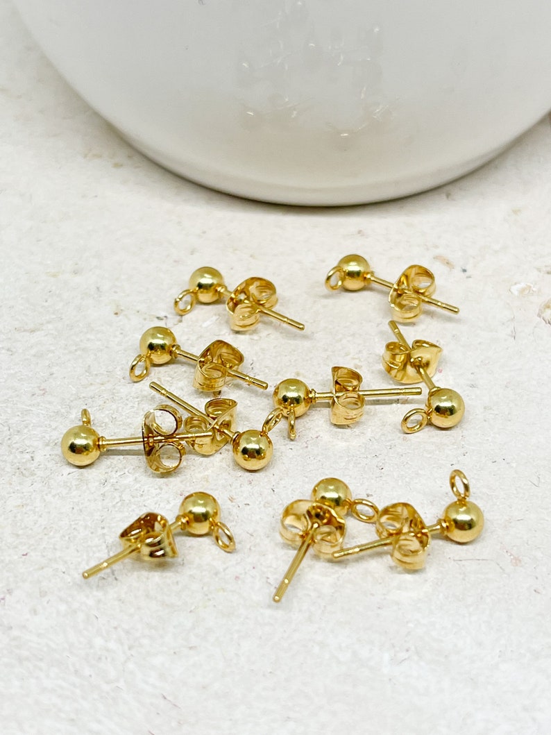 May include: A collection of gold-colored ball stud earring components. Each component features a small, round ball at the front, a post for piercing the ear, and a loop for attaching additional elements. The earrings are displayed on a light-colored surface.