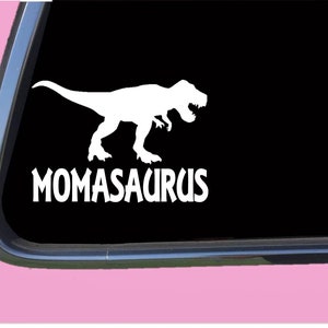 May include: White vinyl decal of a dinosaur silhouette with the text "Momasaurus" on a black background. The decal is on a car window.