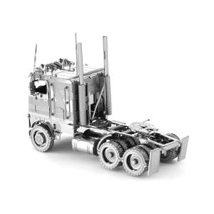 Op de afbeelding: Een zilverkleurig metalen model van een vrachtwagen. Het model heeft gedetailleerde kenmerken, waaronder de cabine, de opleggerkoppeling en meerdere wielen. De vrachtwagen heeft twee lange uitlaatpijpen en lichten op het dak van de cabine.