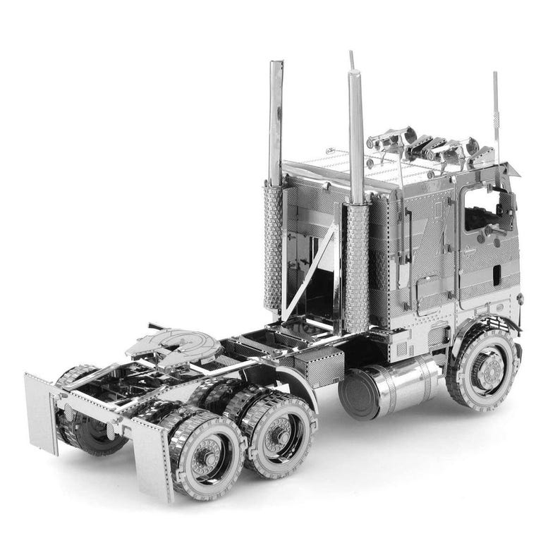 Op de afbeelding: Een zilverkleurig metalen model van een vrachtwagen. De vrachtwagen heeft gedetailleerde componenten, waaronder de cabine, de opleggerkoppeling, de uitlaatpijpen en meerdere wielen. Het model is met precisie geconstrueerd en heeft een reflecterend oppervlak.