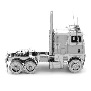 Op de afbeelding: Een zilverkleurig metalen model van een vrachtwagen. Het model heeft gedetailleerde onderdelen, waaronder de cabine, de opleggerkoppeling, de uitlaat en meerdere wielen. Het model staat tegen een witte achtergrond.