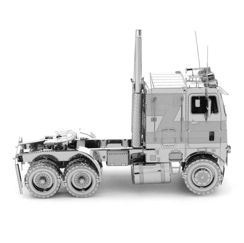 Op de afbeelding: Een zilverkleurig metalen model van een vrachtwagen. Het model heeft gedetailleerde onderdelen, waaronder de cabine, de opleggerkoppeling, de uitlaat en meerdere wielen. Het model staat tegen een witte achtergrond.