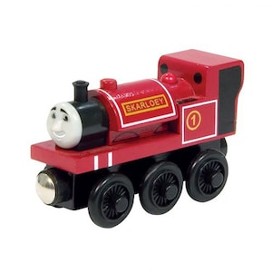 May include: A red wooden toy train engine with a black face, black wheels, and a black chimney. The engine has a white face with a smiling expression and black eyes. The engine has a white stripe on each side and the words "SKARLOEY" printed on the side. The engine has a number "1" printed on the side.
