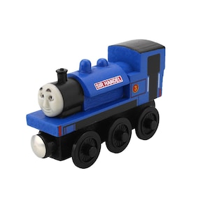 Pode incluir: Um comboio de brinquedo de madeira azul com rodas pretas e um telhado preto. O comboio tem uma cara sorridente e o nome "SIR HANDEL" na lateral. O número "3" também é visível. Brinquedo infantil.