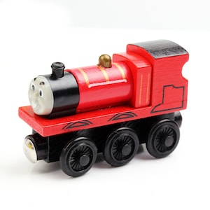 May include: A red wooden toy train engine with black wheels and a black face with white eyes and a smile. The engine has a gold chimney and gold stripes on the side.