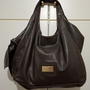 May include: A brown leather handbag with a single shoulder strap. The bag has a slouchy design and a rectangular gold-toned plaque with the brand name "Valentino" on the front. The bag is hanging on a white door.