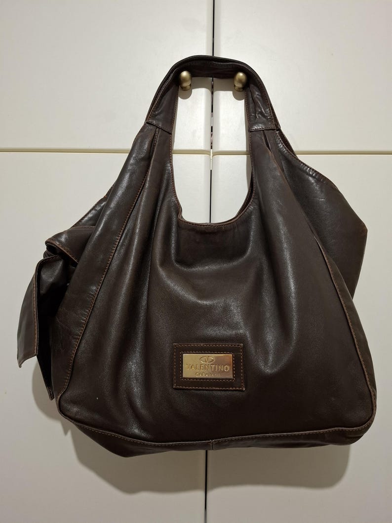 May include: A brown leather handbag with a single shoulder strap. The bag has a slouchy design and a rectangular gold-toned plaque with the brand name "Valentino" on the front. The bag is hanging on a white door.