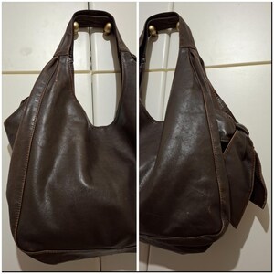 May include: A dark brown leather shoulder bag with a large, open design. The bag has a single strap and a side pocket. The leather appears worn, with visible stitching and a soft, slouchy shape. The bag is hanging on a white door.