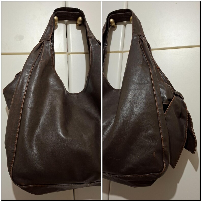 May include: A dark brown leather shoulder bag with a large, open design. The bag has a single strap and a side pocket. The leather appears worn, with visible stitching and a soft, slouchy shape. The bag is hanging on a white door.