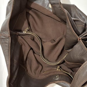 May include: A brown leather tote bag with a soft, slouchy design. The bag features a large interior with a zippered pocket and a brown fabric lining. The exterior has a knotted detail on the side and a snap closure.