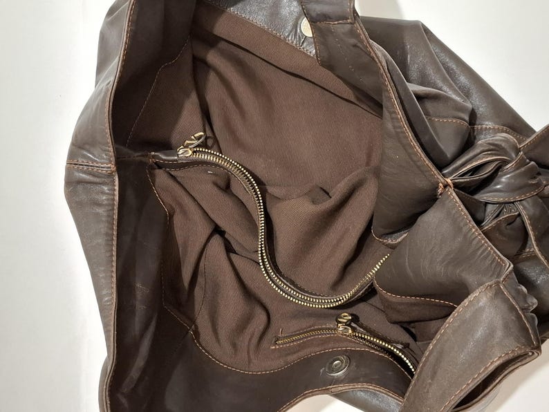 May include: A brown leather tote bag with a soft, slouchy design. The bag features a large interior with a zippered pocket and a brown fabric lining. The exterior has a knotted detail on the side and a snap closure.
