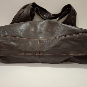 May include: A dark brown leather handbag with a wide, curved shoulder strap. The bag has a soft, slouchy shape with visible stitching details. The leather appears distressed, giving it a vintage look. The bag is designed for everyday use.