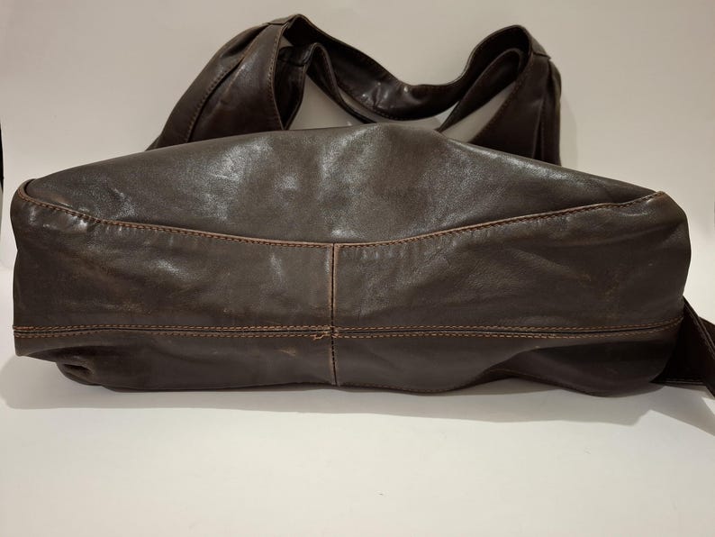 May include: A dark brown leather handbag with a wide, curved shoulder strap. The bag has a soft, slouchy shape with visible stitching details. The leather appears distressed, giving it a vintage look. The bag is designed for everyday use.