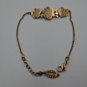 May include: A gold-toned bracelet featuring three flower-shaped charms. The bracelet has a delicate chain and a lobster clasp closure. The brand name "KENZO" is visible on a small tag near the clasp.