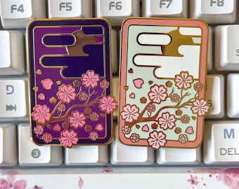 May include: Two enamel pins with a gold finish. The pins feature a branch of pink cherry blossoms with a background of purple and green. The pins are rectangular and have a gold border.