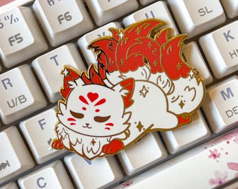 May include: A gold-colored enamel pin featuring a white fox with red and gold accents. The fox has a curled tail and is lying down with its head resting on its paws. The pin is on a white keyboard.