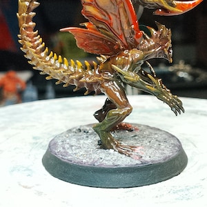 May include: A detailed miniature figurine of a fantasy creature with a reptilian body, insect-like wings, and a curved tail. The figure is painted in shades of brown, green, and orange, and stands on a circular base. The wings are a vibrant orange.