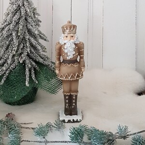 Könnte beinhalten: Dekorative Nussknacker-Figur in Braun-, Gold- und Weißtönen. Die Figur trägt eine Krone und Stiefel und hat einen weißen Bart. Im Hintergrund befindet sich ein kleiner, schneebedeckter Weihnachtsbaum.