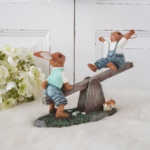 Könnte beinhalten: Eine skurrile Figur zeigt zwei braune Hasen, die auf einer Holzwippe spielen. Ein Hase trägt eine türkisfarbene Weste und eine Jeans-Latzhose, der andere ein weißes Hemd und eine Jeans-Latzhose. Der Sockel zeigt Gras und Pilze, im Hintergrund ein Kranz.