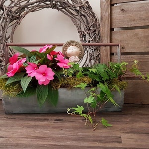 Könnte beinhalten: Eine rechteckige Metall-Pflanzschale gefüllt mit leuchtend rosa Blüten, grünem Laub und Moos. Eine kleine Figur befindet sich zwischen den Pflanzen. Der Pflanzkasten hat einen Metallgriff und steht auf einer Holzoberfläche, mit einem geflochtenen Kranz im Hintergrund.